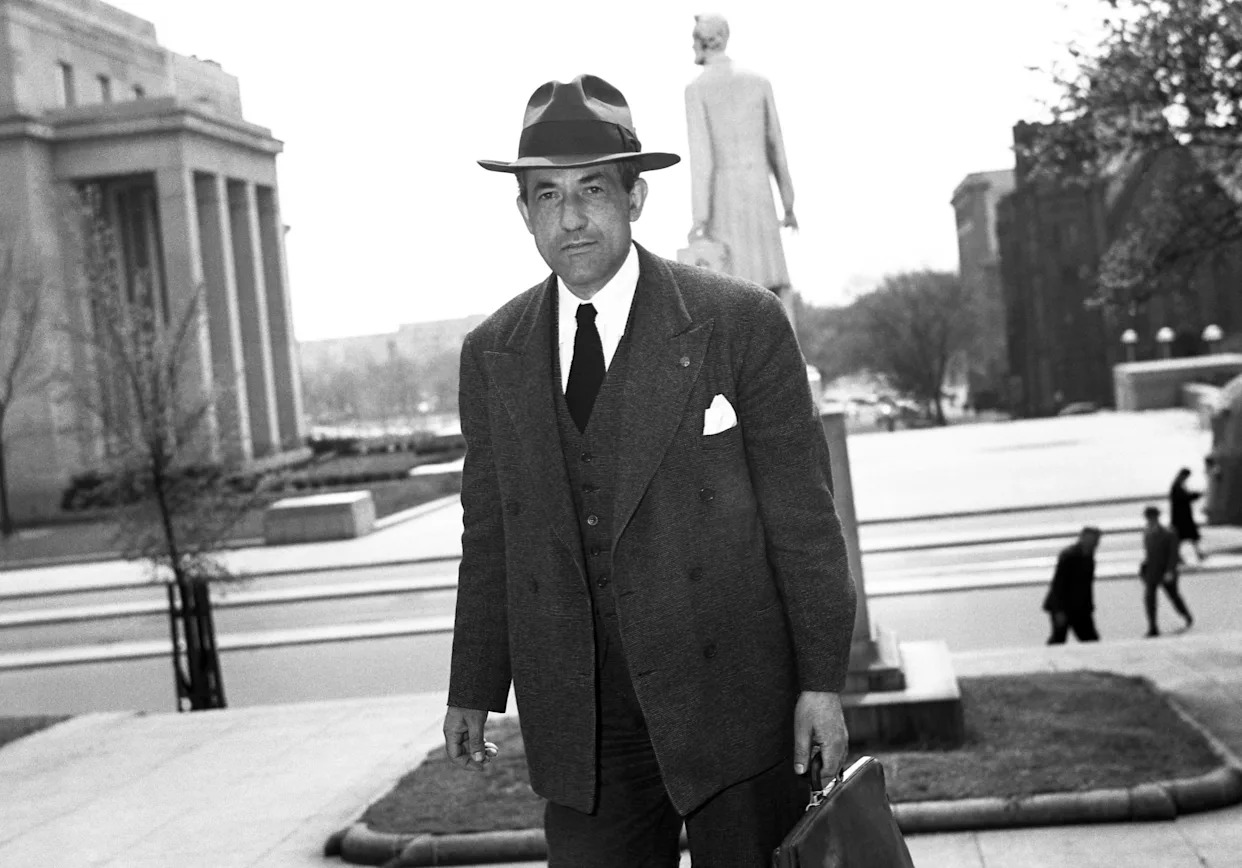 A black-and-white photo of a man in a hat, suit, and tie walking across a city street, with marble columns and a statue behind him.