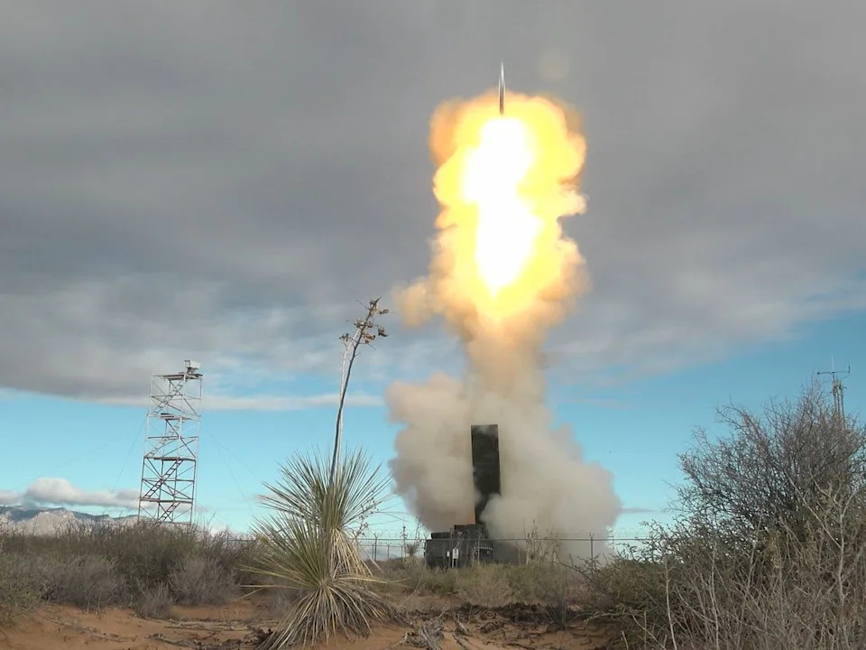 A missile being fired from a launcher in a desert landscape with gray clouds across a blue sky in the background.