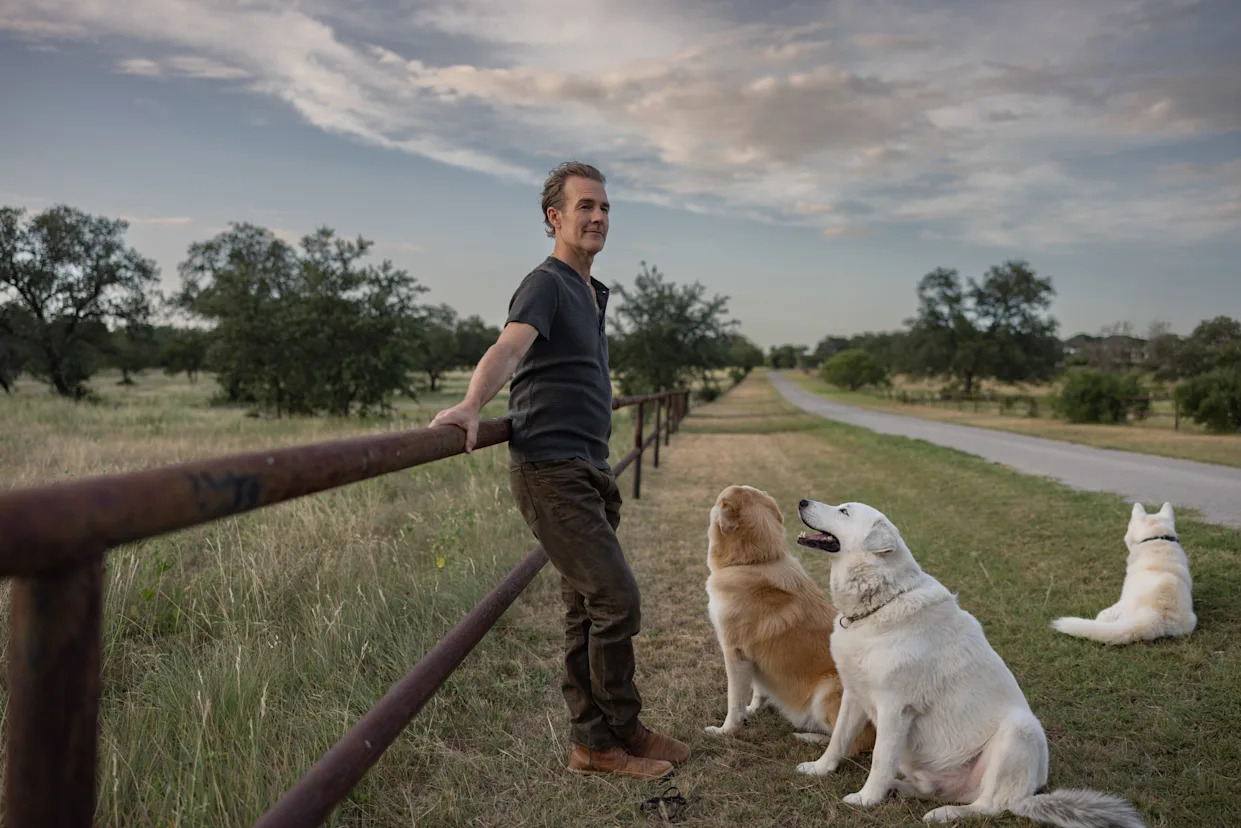 James Van Der Beek leaning against a fence near an open road with three dogs