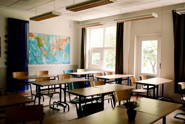 Empty classroom with desks, chairs, a world map on the wall, and large windows letting in natural light