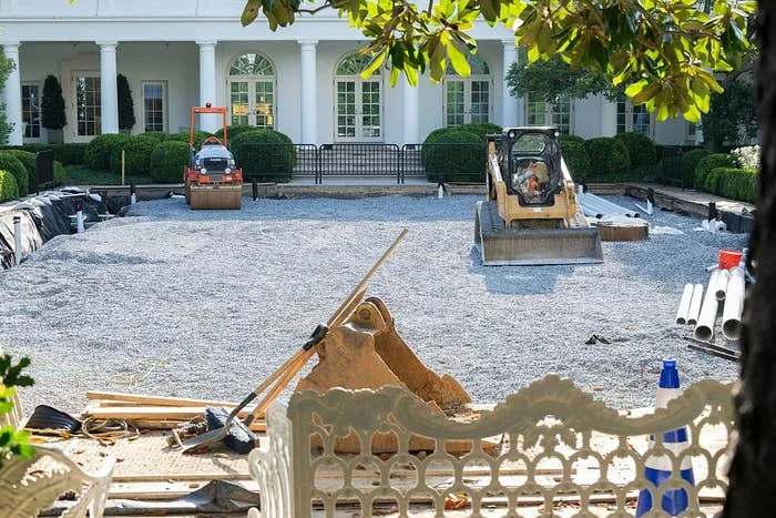 A construction site with heavy machinery preparing the ground in a garden setting outside a large white building