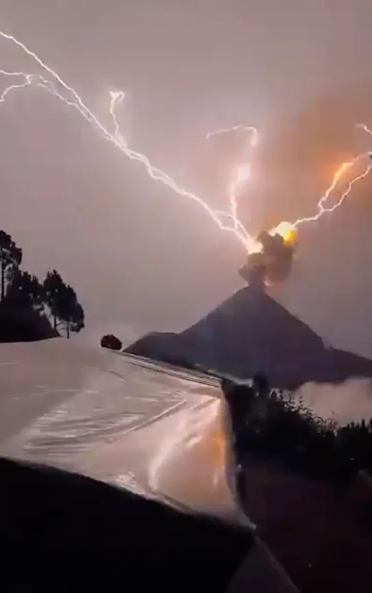 Lightning strikes near erupting volcano, with ash and smoke billowing. A tent is visible in the foreground among silhouetted trees