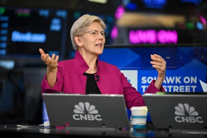 A speaker gestures while sitting at a CNBC news desk, engaging in a discussion, with stock market displays in the background