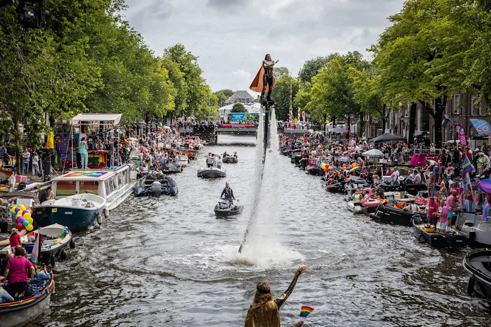 Festival-goers in many boats watch a person performing on a flyboard in a canal.