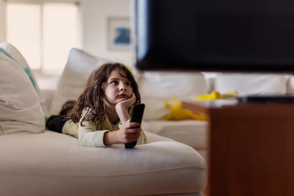 Young child lying on a couch, holding a TV remote, gazing towards the television with a thoughtful expression