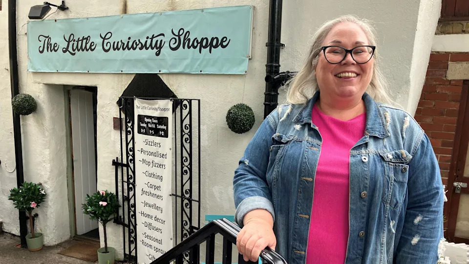A blond, bespectacled woman stands in front of a shop. She is wearing a pink top and denim jacket over it as she holds on to a railing. A banner above the shop entry reads "The Little Curiosity Shoppe".