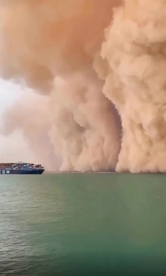 A large dust storm approaches a massive cargo ship at sea, creating a stark contrast with the water's calm surface