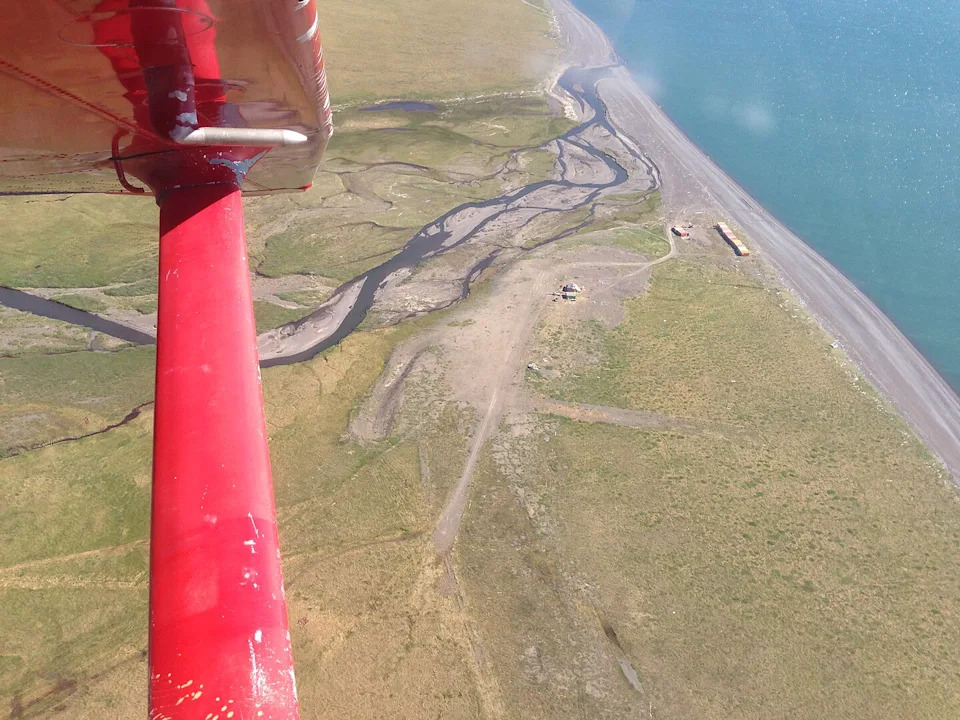 An aerial view, partially obscured by a red plane wing and strut on the left, shows a rugged coastline where a meandering river meets a large body of water. The landscape is a mix of green tundra and dirt paths, with visible signs of human activity, including some structures or equipment on the narrow strip of land between the river and the sea. The water appears blue-green, and the sky is bright.