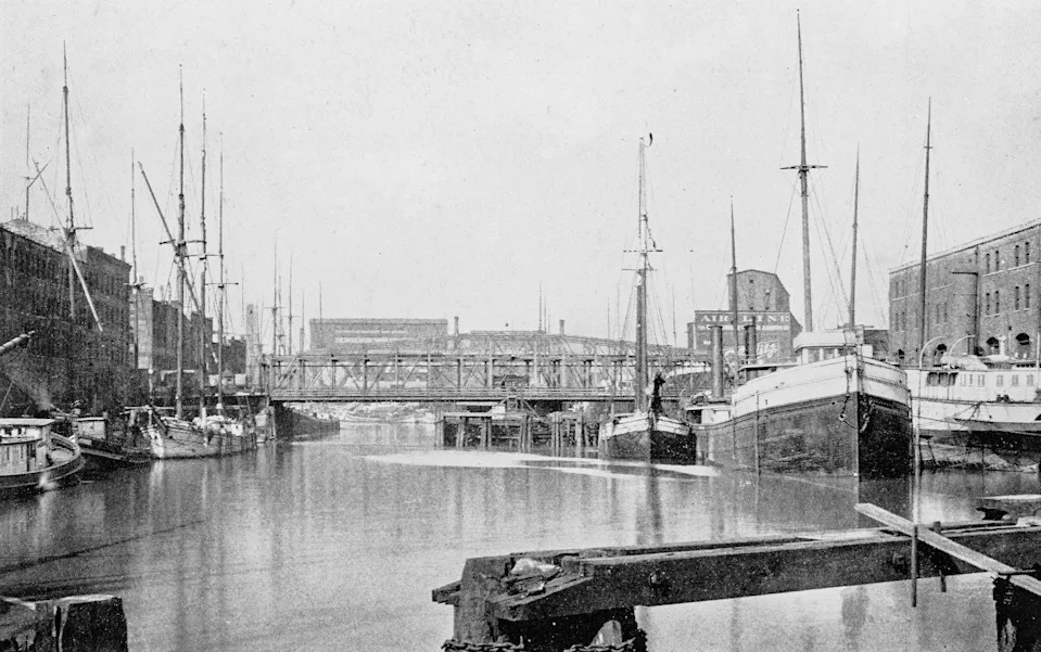 Historic harbor scene with several docked sailboats and warehouses, featuring an industrial bridge in the background