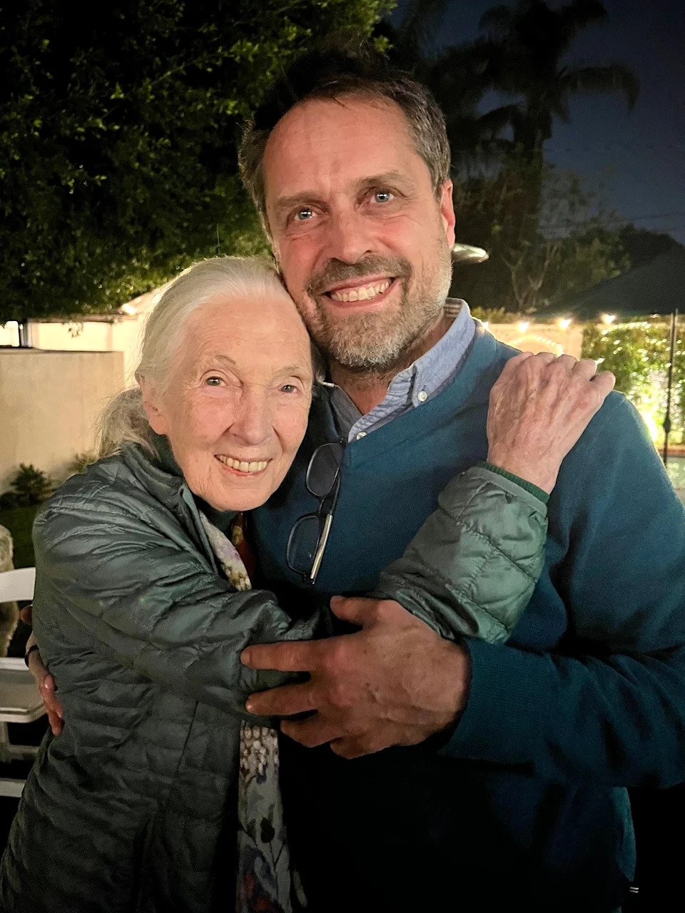 Two people smiling and hugging outdoors at night, one wearing a quilted jacket and the other in a sweater with glasses hanging from the collar