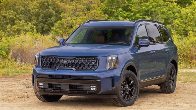 a 2/3rds view of a 2025 Kia Telluride in blue parked on dirt with green trees in the background