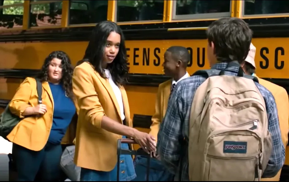 Group of students in yellow blazers talking by a school bus, one extends a hand to another