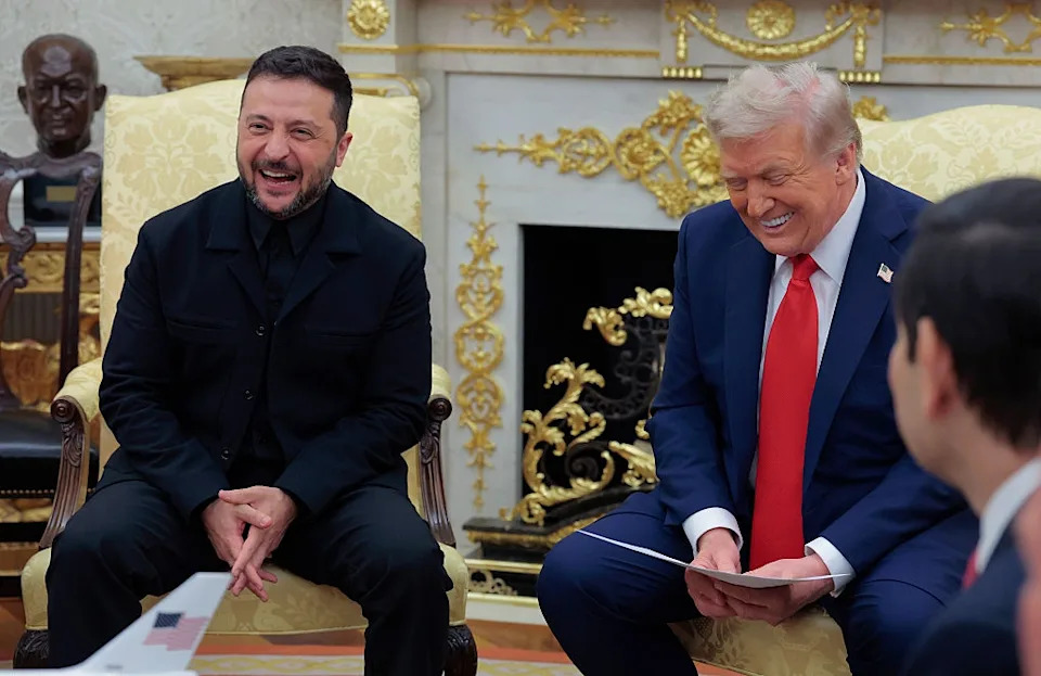 Two men seated and laughing in an ornate room with decorative elements and a fireplace